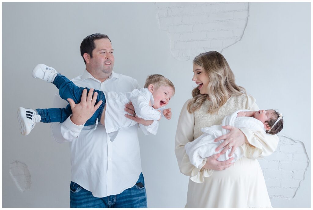 dad is playing with toddler boy while mom is laughing and holding her newborn baby girl - Newborn Photographer in Shreveport