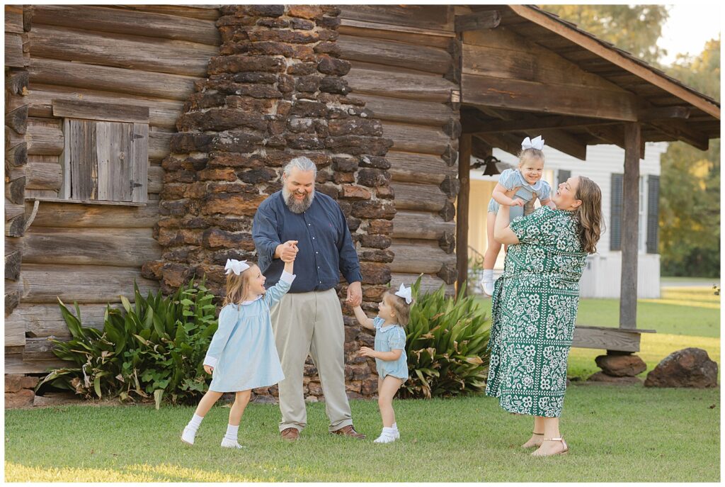 Relaxed family photo session in Bossier with parents and three daughters in front of rustic cabin, wearing blue and green outfits